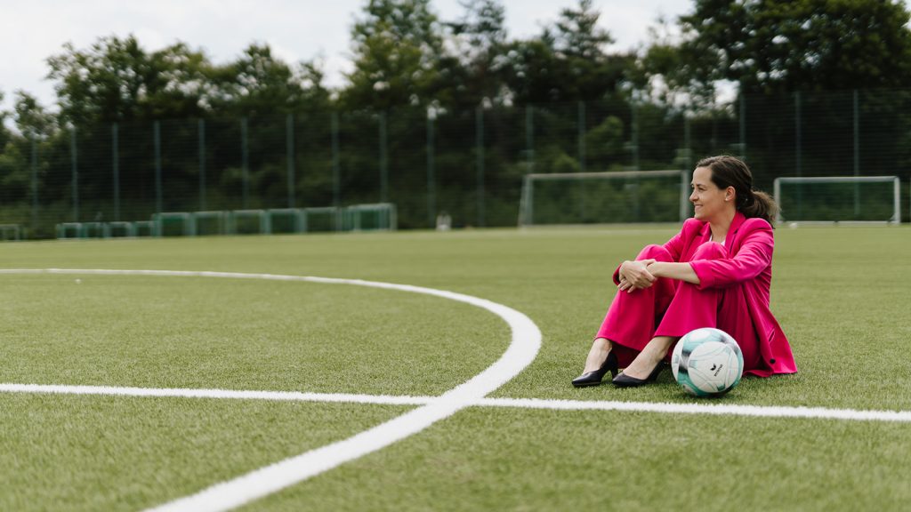 Julia Wiedemann Expertin für Sport-Sponsoring Julia sitzt mit Anzug auf Fußballplatz